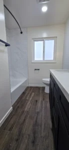Bathroom remodel in Oakland CA featuring white tub surround, dark vanity, and wood-look tile flooring